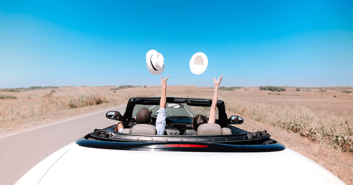 Two people in a convertible automobile on the road throwing up their hats